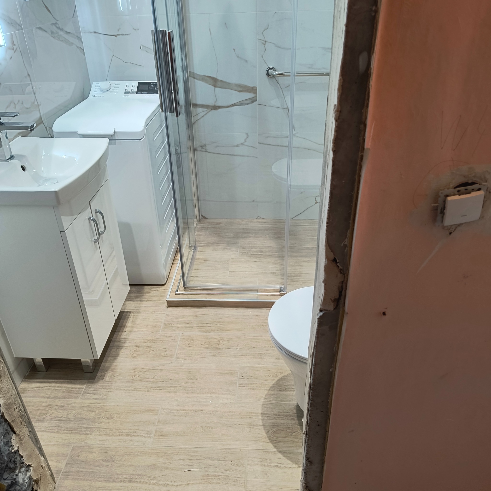 Wykończona łazienka z kabiną prysznicową, umywalką z szafką i pralką. Jasne płytki imitujące drewno na podłodze, marmurowe na ścianach. Częściowo widoczna ściana w trakcie prac.