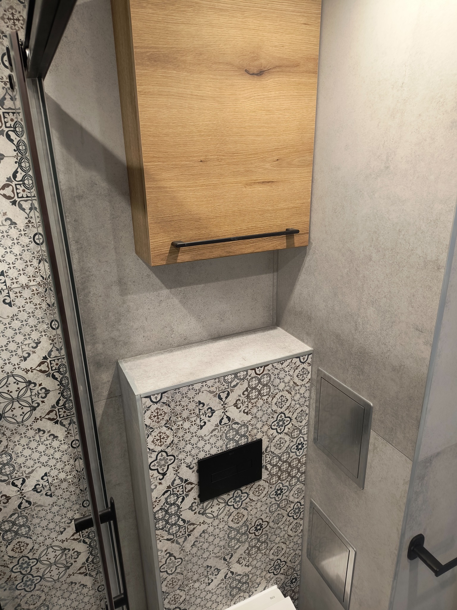 Wykończona łazienka: kabina prysznicowa z wzorzystymi płytkami, drewniana szafka nad zabudową toalety z czarnym przyciskiem, szare ściany i metalowe rewizje.