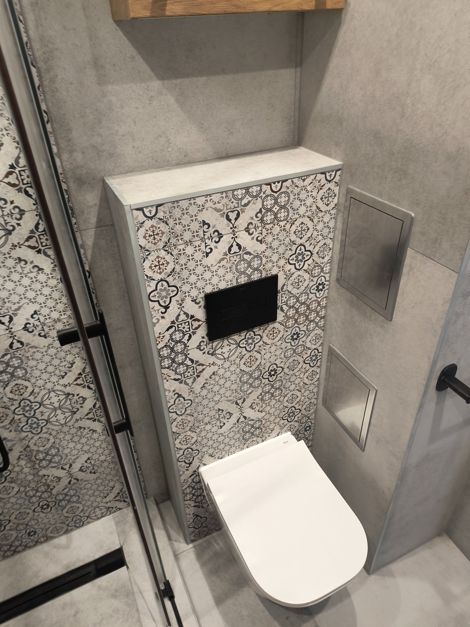 Nowoczesna łazienka z podwieszaną toaletą i obudową w dekoracyjne płytki, szara ściana, metalowe rewizje. Minimalistyczny design z akcentem wzorzystych płytek.