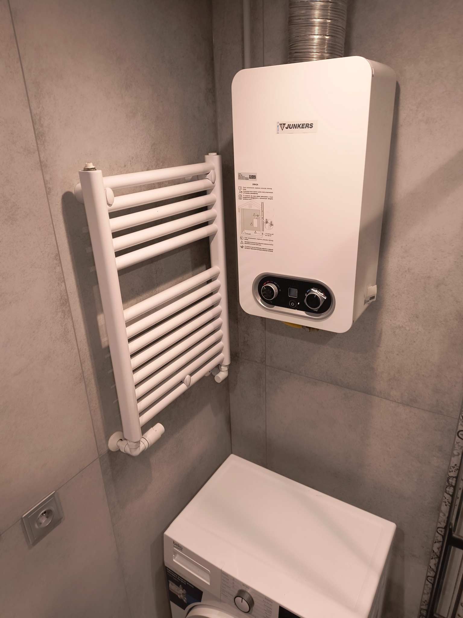 Szary wnętrze łazienki z grzejnikiem drabinkowym, wiszącym piecem gazowym Junkers i pralką Beko. Minimalistyczny design, płytki imitujące beton.