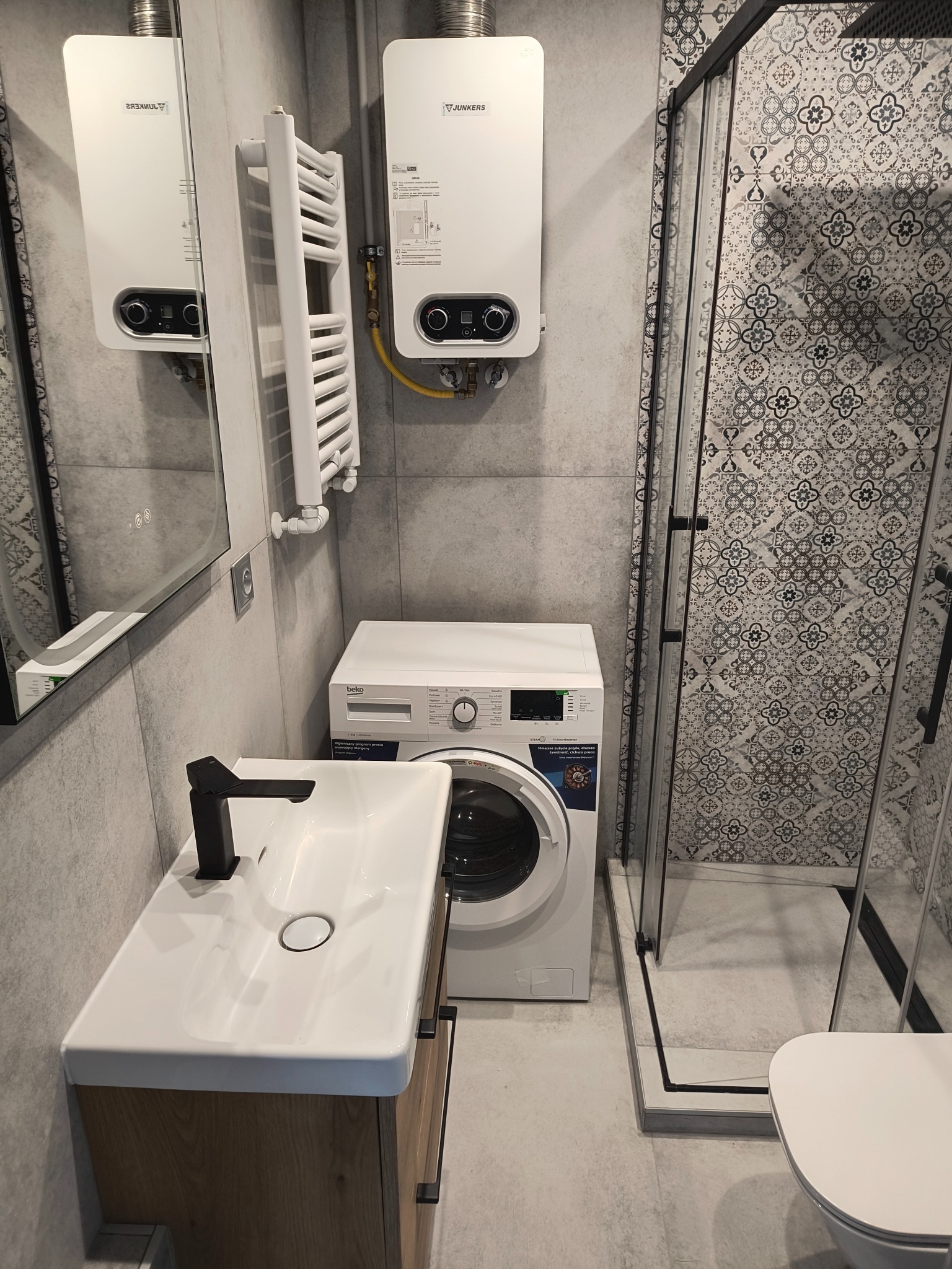 Nowoczesna łazienka z kabiną prysznicową, umywalką z szafką, pralką i grzejnikiem. Szare płytki i wzorzyste płytki w kabinie tworzą spójną całość.