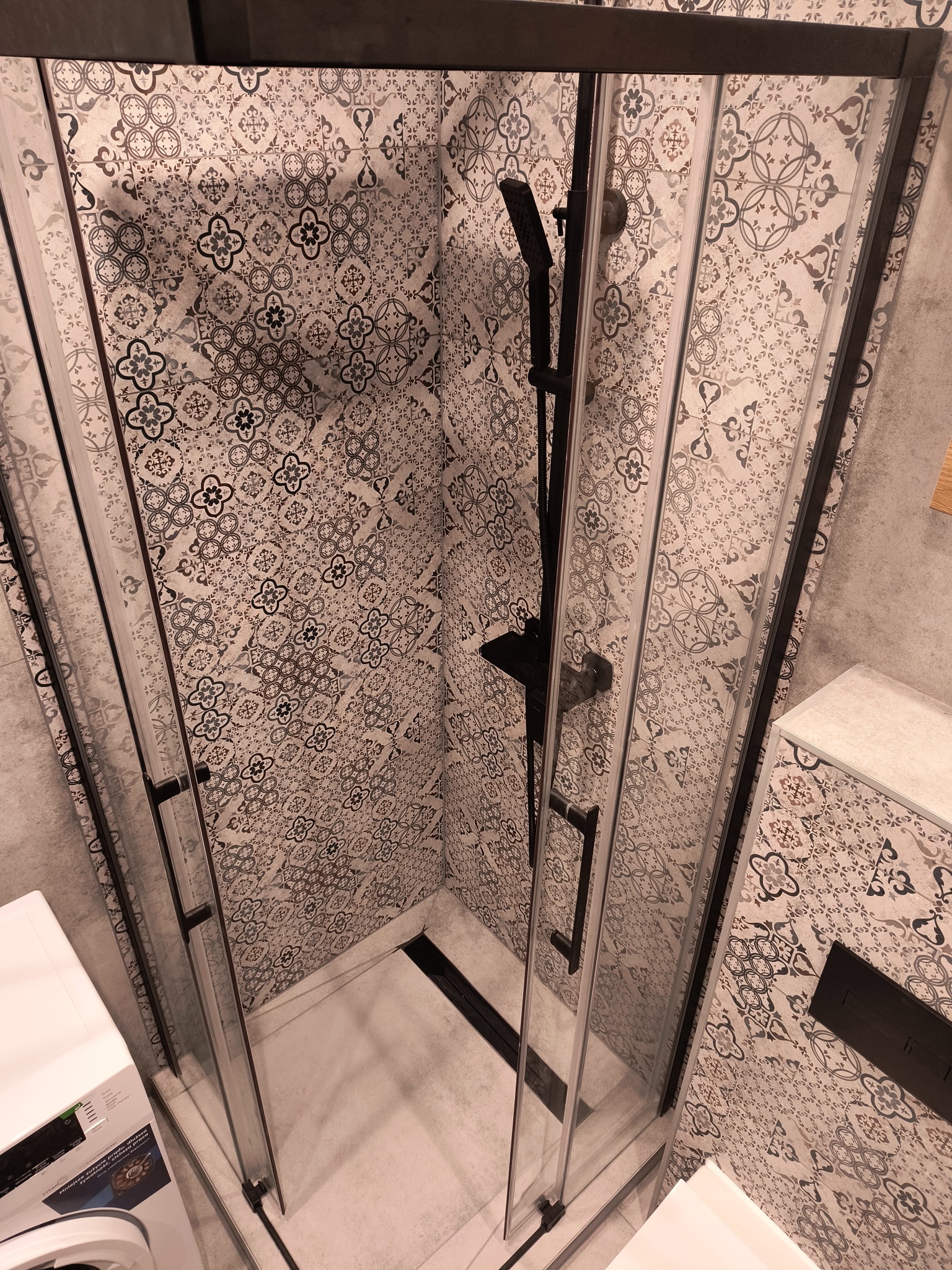 Kabina prysznicowa z czarnymi profilami i deszczownicą na tle wzorzystych płytek w odcieniach szarości. Widoczna pralka i fragment umywalki.