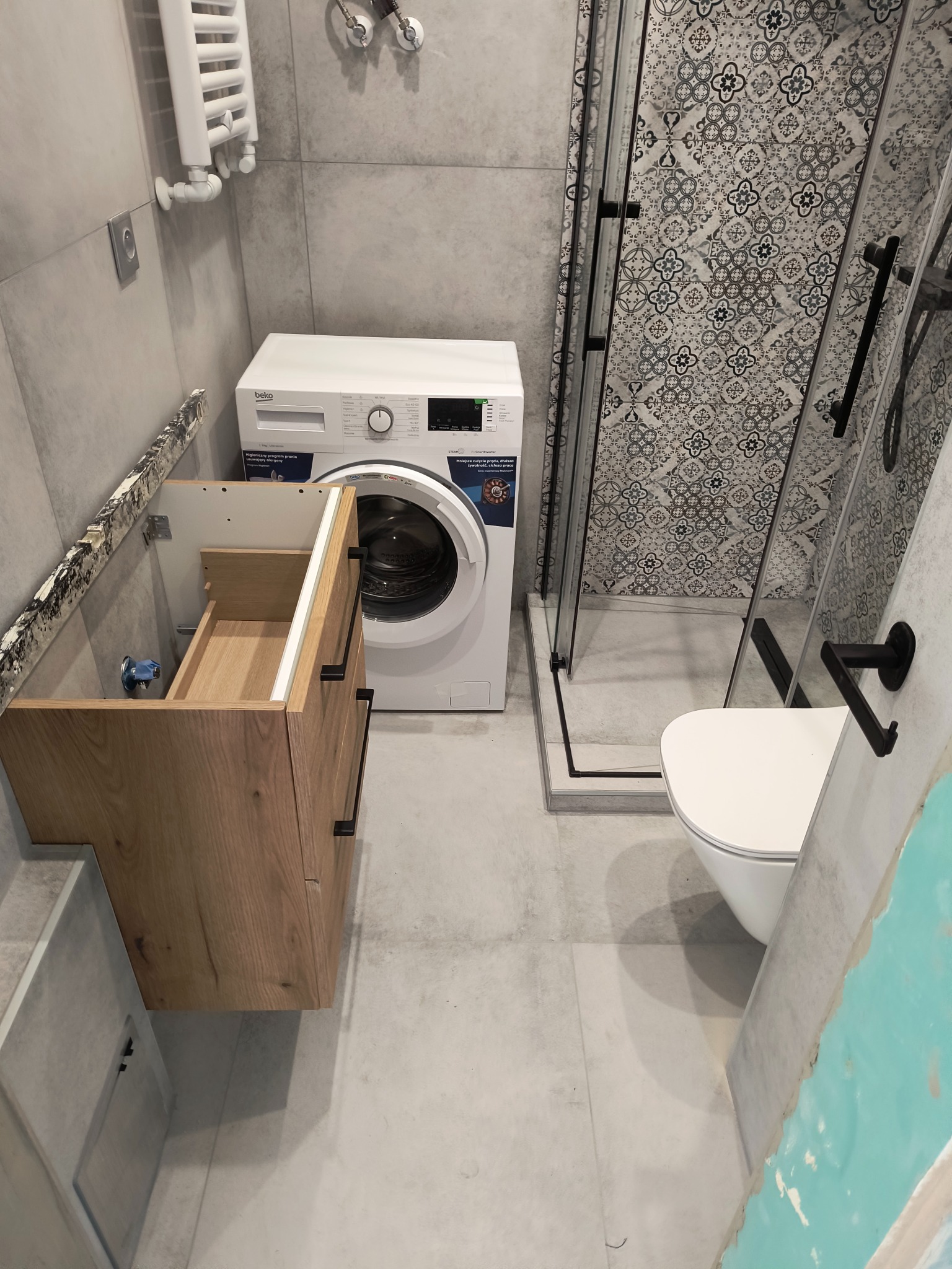 Jasna łazienka po remoncie: kabina prysznicowa z wzorzystymi płytkami, umywalka z szafką z drewna, biała pralka i toaleta. Minimalistyczny design.