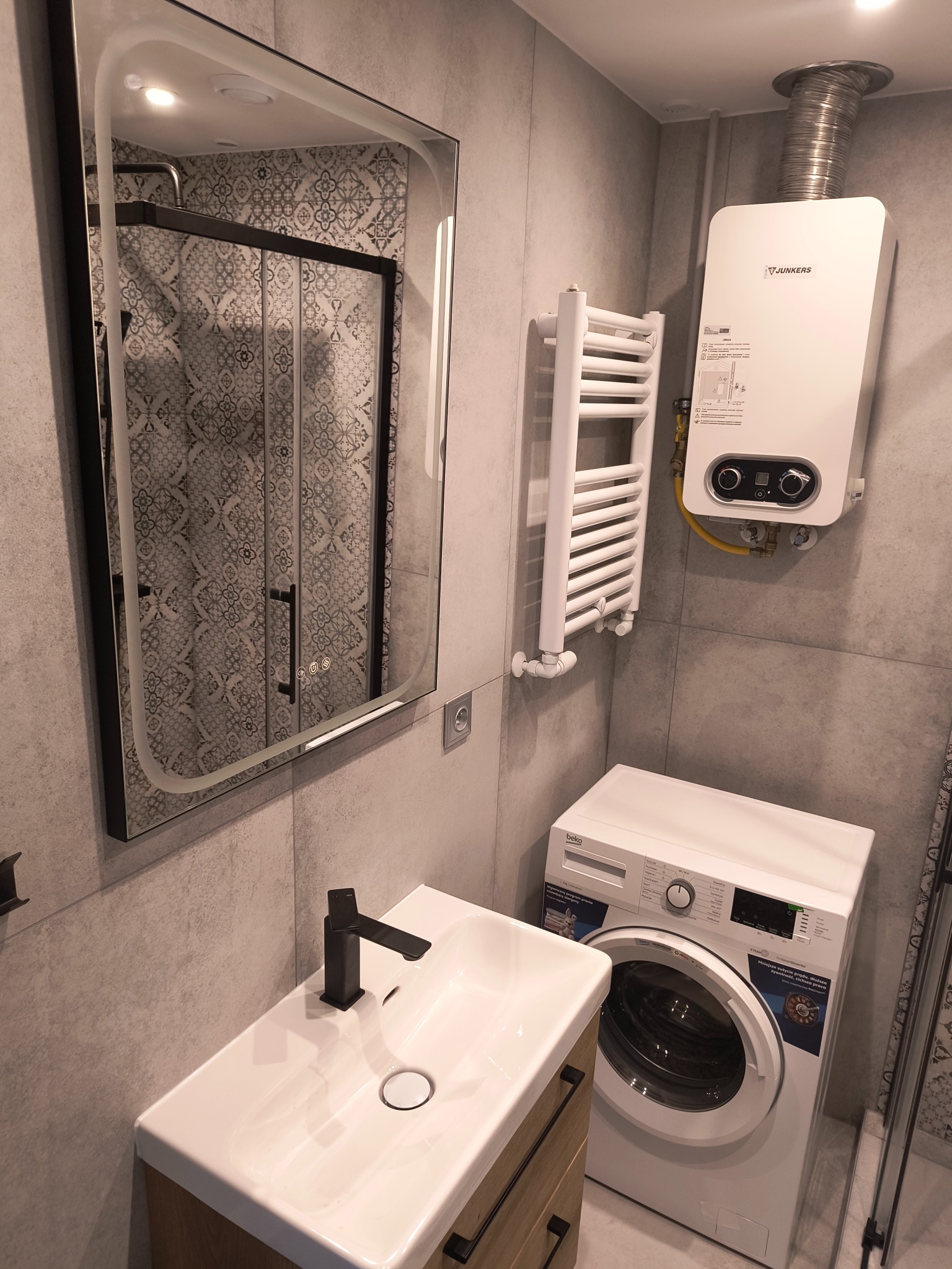 Nowoczesna łazienka: umywalka z szafką, lustro z podświetleniem, pralka Beko i bojler Junkers na szarej ścianie. Kabina prysznicowa z wzorzystymi płytkami w odbiciu lustra.