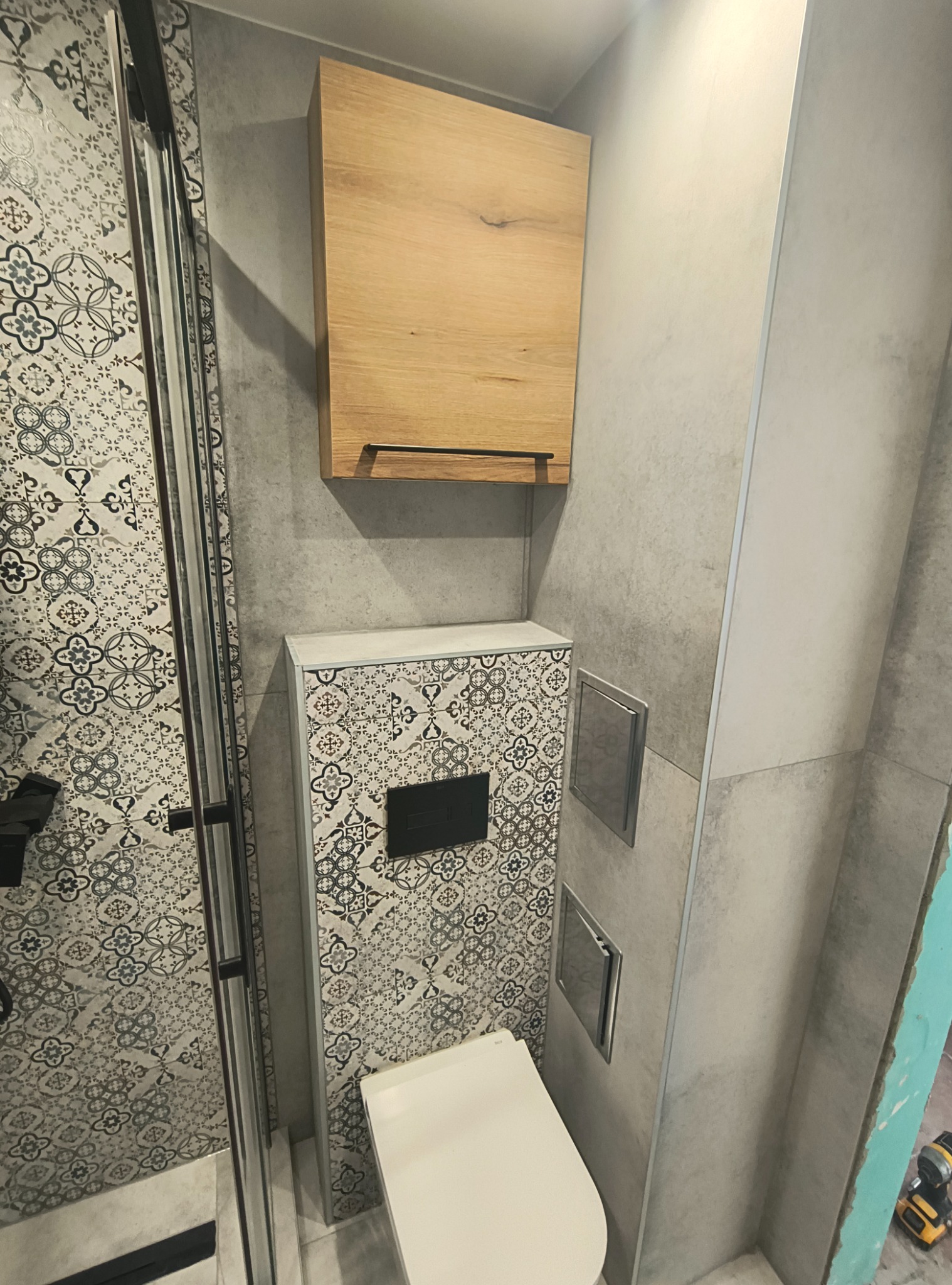 Wykończona łazienka z kabiną prysznicową, toaletą podwieszaną w zabudowie z wzorzystymi płytkami i szafką wiszącą z drewna. Szare płytki na ścianach.