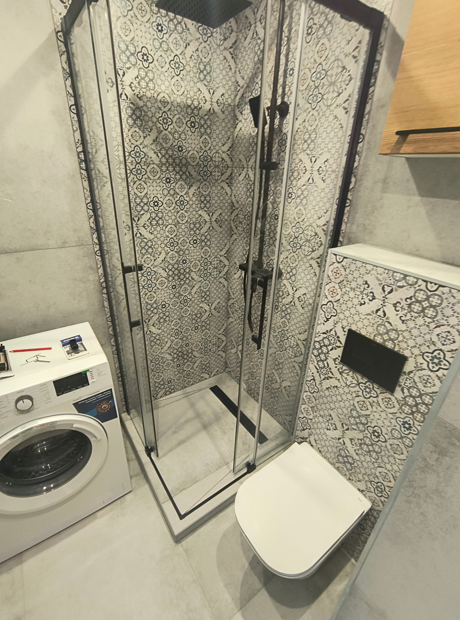 Wykończona łazienka z kabiną prysznicową typu walk-in z czarnymi profilami, toaletą i pralką. Wzorzyste płytki na ścianach i podłodze.