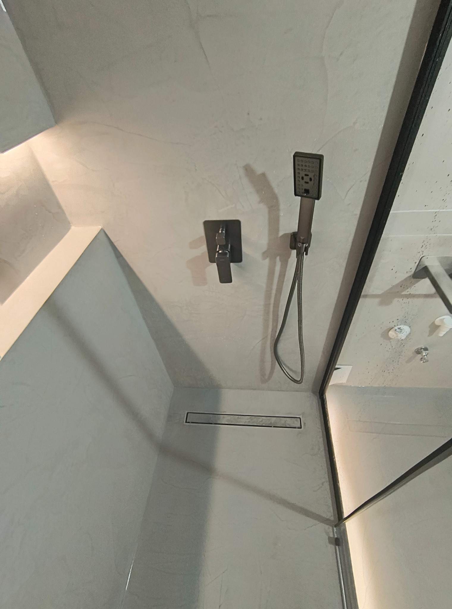 Minimalistyczna łazienka z szarym mikrocementem na ścianach, czarną armaturą i odpływem liniowym. Kabina prysznicowa z czarnymi profilami.