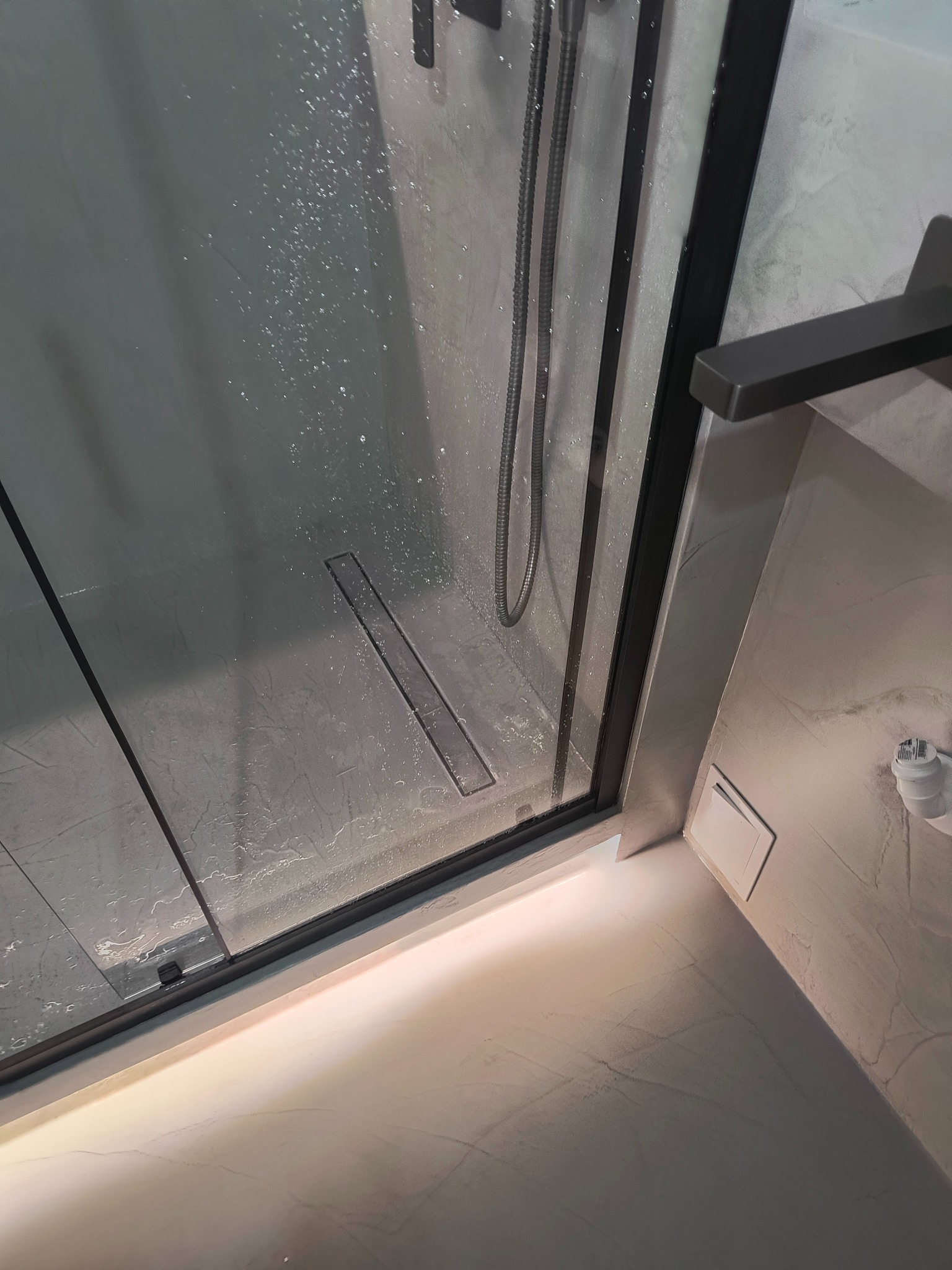 Nowoczesna kabina prysznicowa z odpływem liniowym i deszczownicą, wykończona w szarościach. Widoczne detale: czarne ramy, minimalistyczny design, oświetlenie podłogowe.