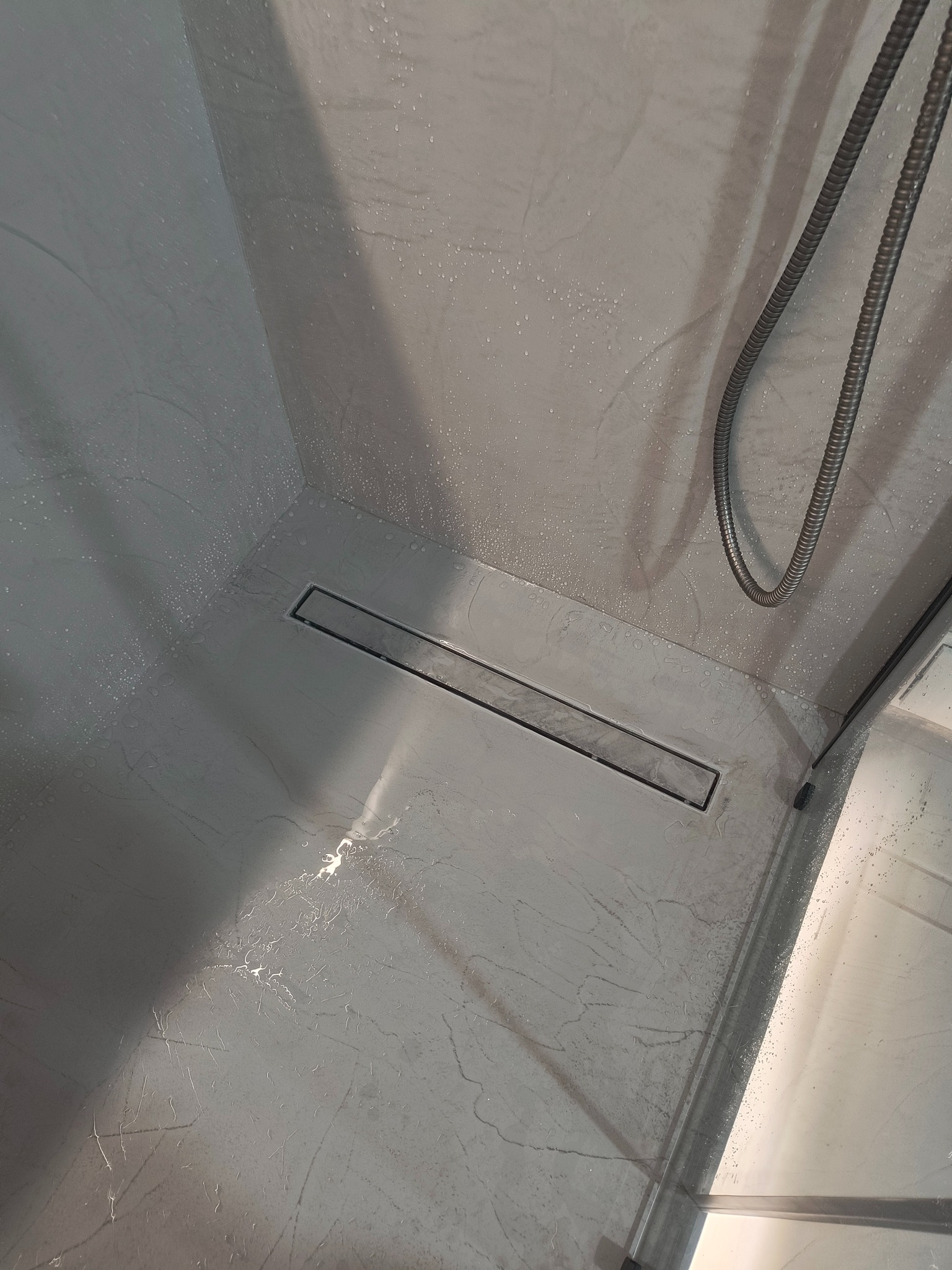 Szara kabina prysznicowa z odpływem liniowym i śladami wody, wykończona w stylu imitującym marmur. Widoczny wąż prysznicowy i fragment szklanej ścianki.