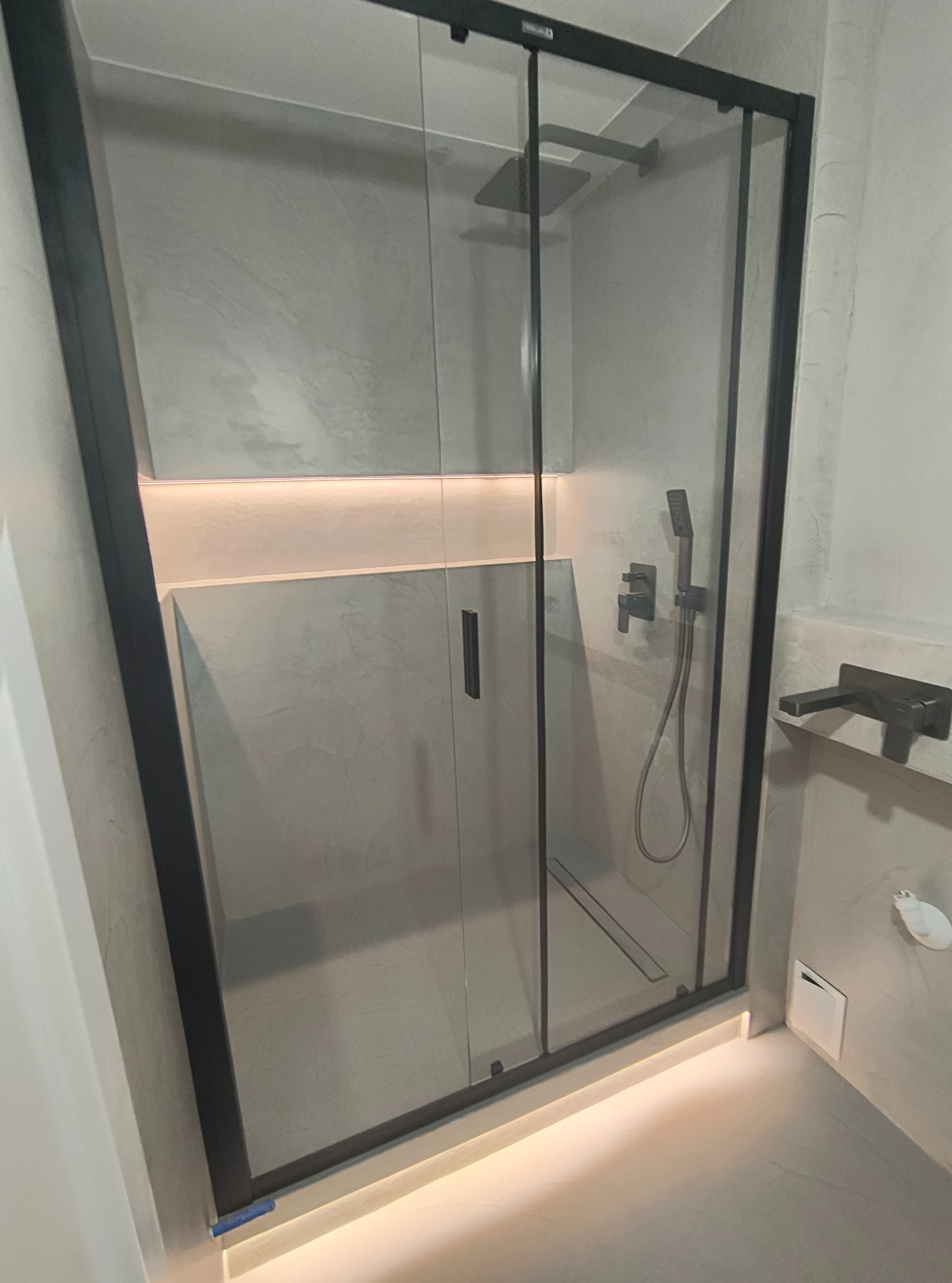 Nowoczesna kabina prysznicowa z czarnymi profilami i deszczownicą, wykończona szarym mikrocementem, z subtelnym oświetleniem LED u dołu i wnęce. Minimalistyczny design.