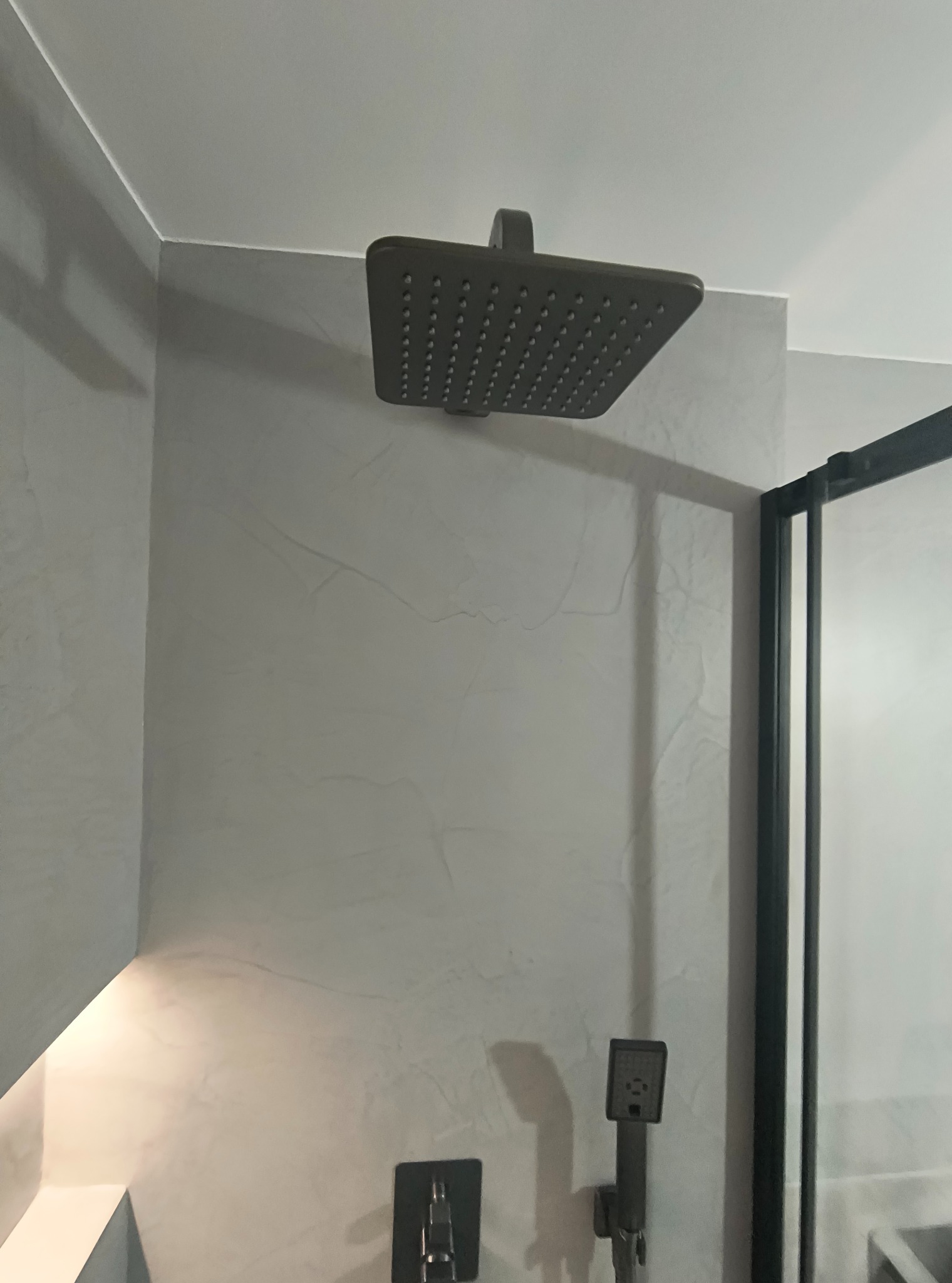 Nowoczesna łazienka z szarą ścianą wykończoną mikrocementem, deszczownicą i czarną kabiną prysznicową. Minimalistyczny design.