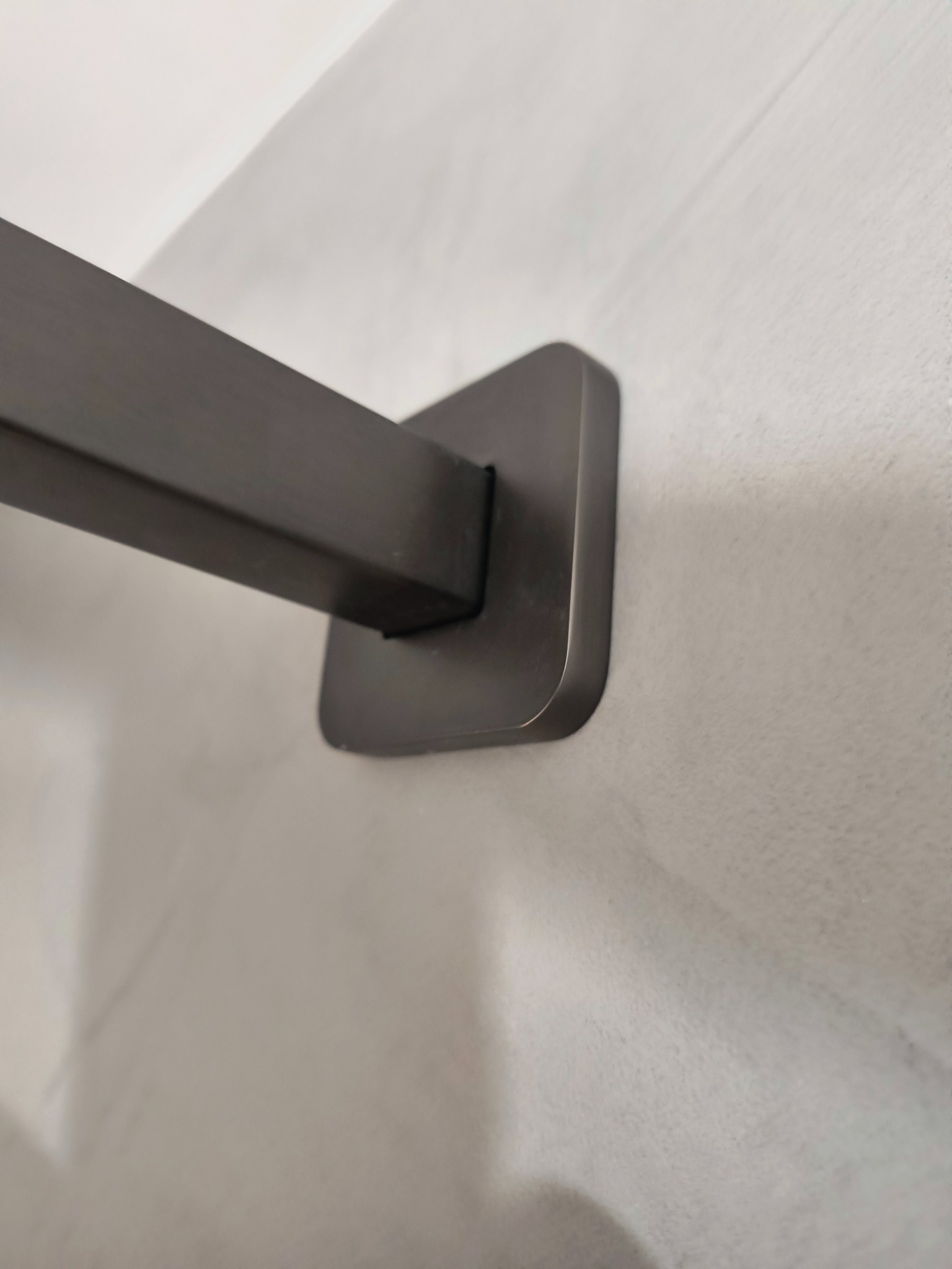 Minimalistyczny, grafitowy uchwyt na ręcznik, zamontowany na jasnoszarej ścianie z efektem betonu. Nowoczesny design, subtelne oświetlenie.