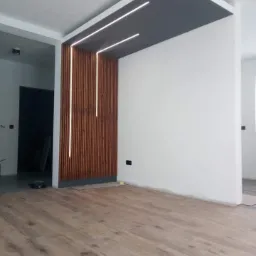 Nowoczesny salon z drewnianą ścianką działową, oświetleniem LED i panelami podłogowymi w odcieniach brązu.