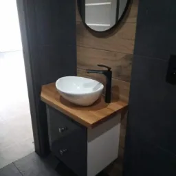 Nowoczesna łazienka z umywalką nablatową na drewnianym blacie, czarną armaturą i okrągłym lustrem na ścianie wyłożonej drewnopodobnymi płytkami.