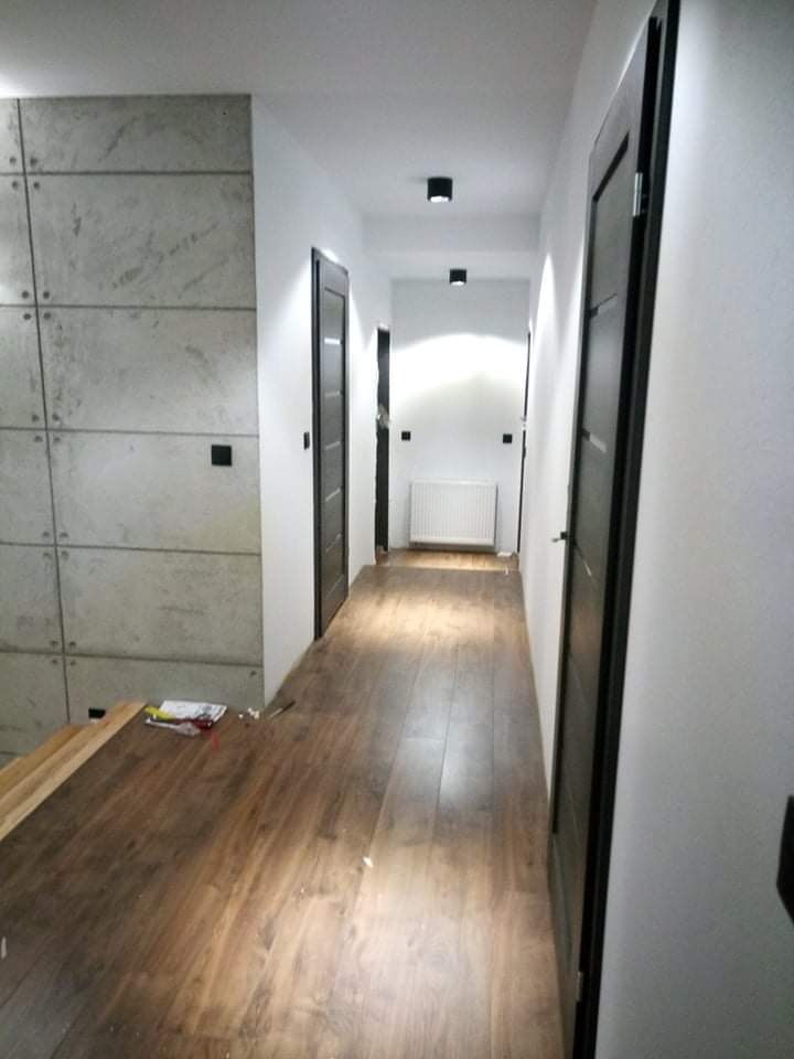 Wnętrze korytarza z panelami imitującymi beton, drewnianą podłogą i czarnymi drzwiami, w tle widoczny grzejnik.