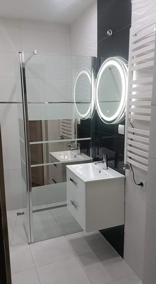 Nowoczesna łazienka z kabiną prysznicową ze szkła, białą szafką pod umywalkę z dwiema szufladami, dwoma okrągłymi lustrami z podświetleniem LED oraz białym grzejnikiem drabinkowym.