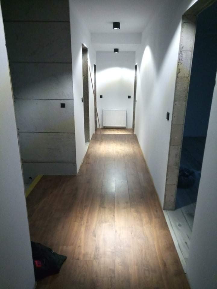 Długi korytarz po remoncie z panelami podłogowymi imitującymi drewno, białe ściany z czarnymi włącznikami, sufit z trzema czarnymi lampami punktowymi, widoczny grzejnik w oddali.