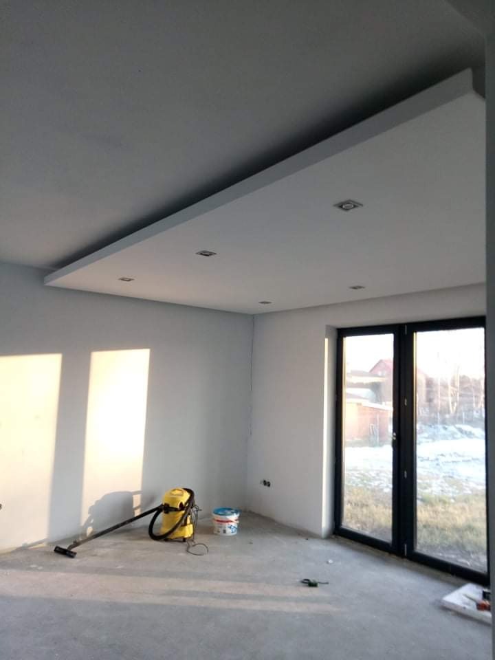 Wnętrze świeżo wykończonego pokoju z pomalowanymi ścianami na biało, widoczne narzędzia sprzątające, sufit podwieszany z oświetleniem punktowym, duże okno z widokiem na zewnątrz.