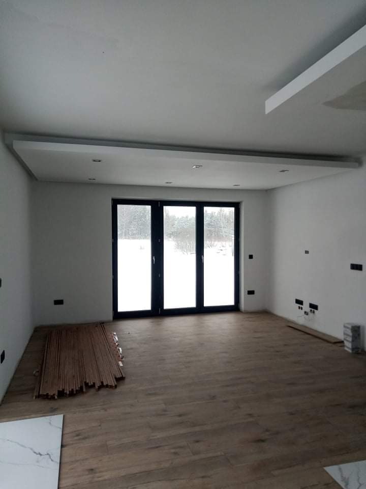 Wnętrze w trakcie remontu: drewniana podłoga ułożona, przy ścianie stos desek podłogowych, widoczne przygotowania do dalszych prac wykończeniowych, duże okno balkonowe z widokiem na zimowy krajobraz.