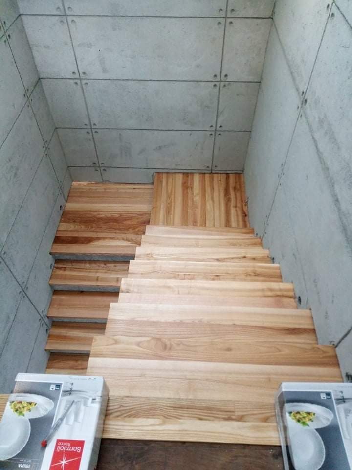 Montaż drewnianych schodów w surowym, betonowym wnętrzu. Widoczne pudełka z zastawą stołową na dole schodów.