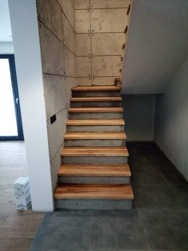 Betonowe schody z drewnianymi stopniami, zamontowane przy ścianie z paneli betonowych, widok z dołu na górę, w tle szara podłoga z płytek.