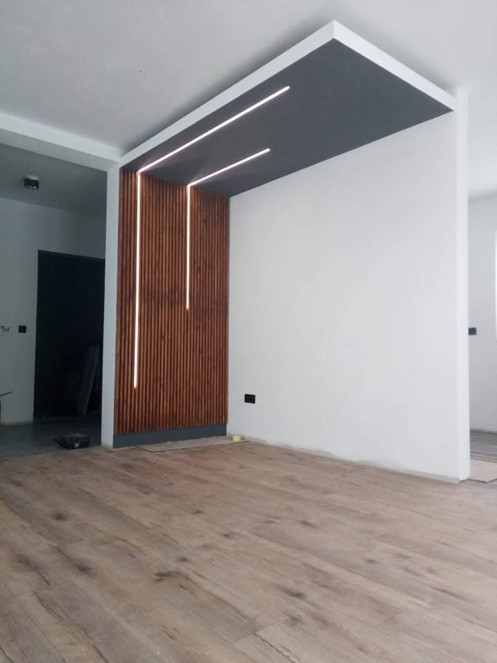 Nowoczesny salon z drewnianą ścianką działową, oświetleniem LED i panelami podłogowymi w odcieniach brązu.