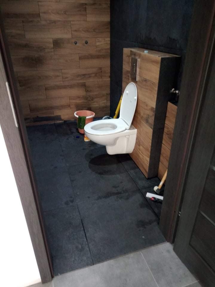 Nowoczesna toaleta z drewnopodobnymi płytkami na ścianach, ciemną podłogą i wiszącą muszlą klozetową, widoczny kubeł i narzędzia na podłodze.