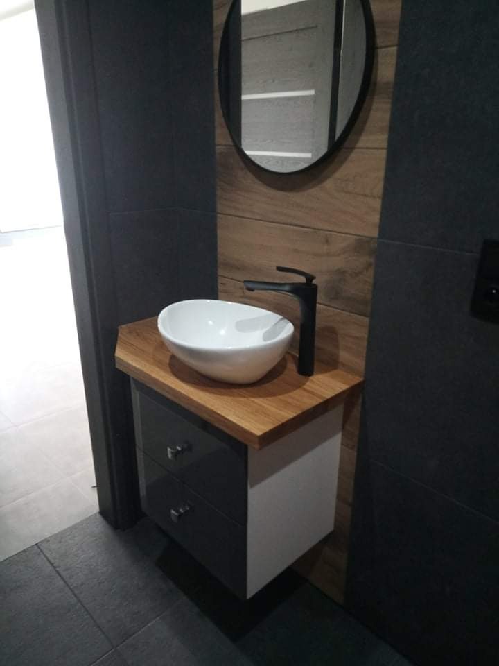 Nowoczesna łazienka z umywalką nablatową na drewnianym blacie, czarną armaturą i okrągłym lustrem na ścianie wyłożonej drewnopodobnymi płytkami.