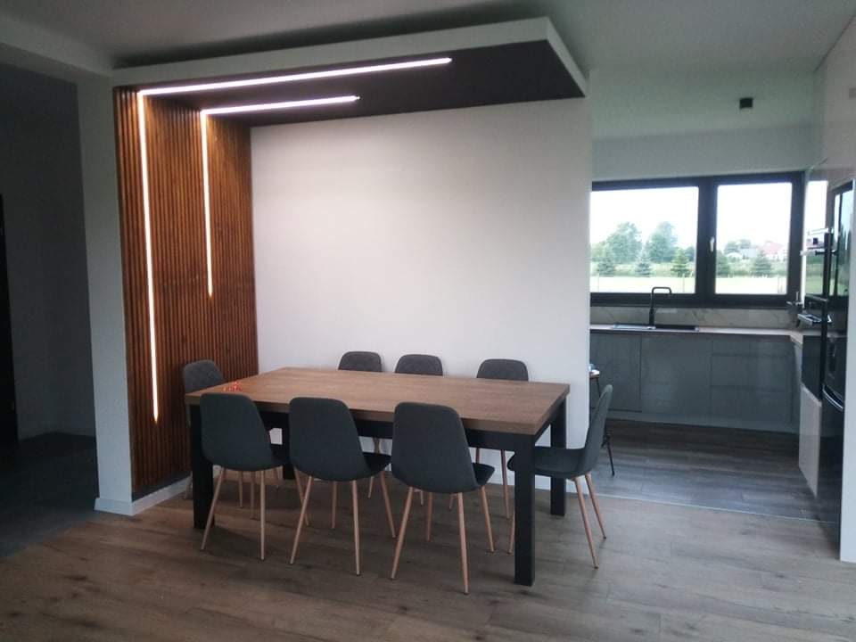 Nowoczesna jadalnia z drewnianą ścianką działową, podświetloną pionowymi lampami LED, oraz stołem z szarymi krzesłami, widok na kuchnię w tle.