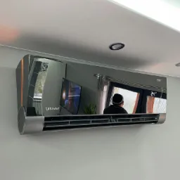 Nowoczesny, srebrny klimatyzator Rotenso zamontowany na ścianie, z widocznym odbiciem wnętrza pokoju, w tym telewizora i osoby w czapce.