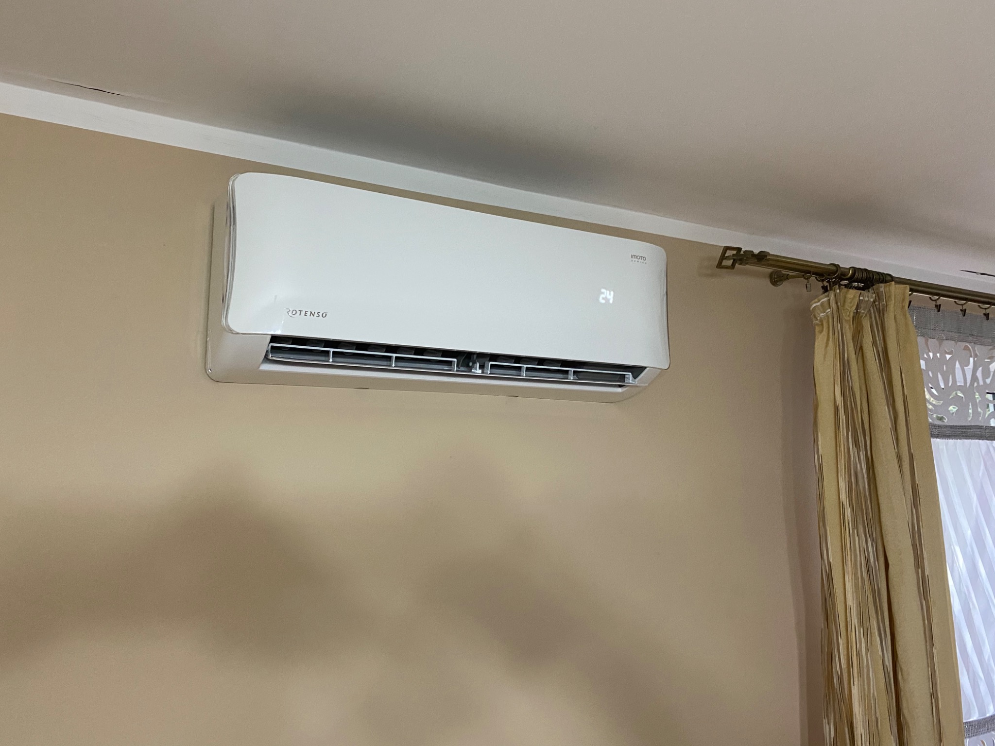 Biały klimatyzator ROTENSO zamontowany na beżowej ścianie w pomieszczeniu z zasłoniętym oknem, wyświetlający temperaturę 24 stopnie.