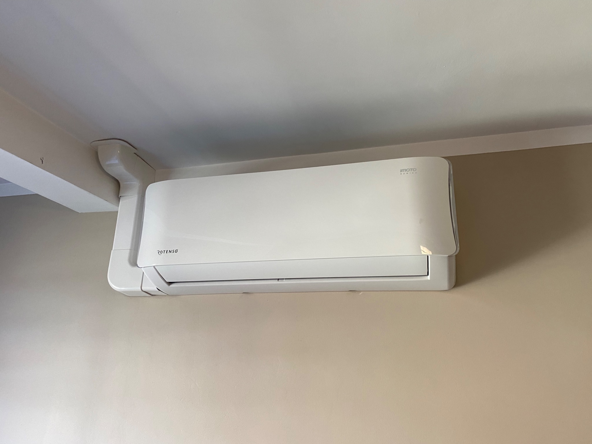Biała jednostka wewnętrzna klimatyzacji marki Rotenso, zamontowana na beżowej ścianie w pomieszczeniu z białym sufitem.
