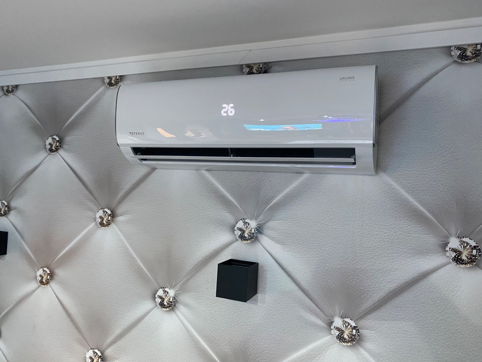 Biały klimatyzator z wyświetlaczem temperatury 26 stopni, zamontowany na ścianie wykończonej pikowaną tapicerką z ozdobnymi kryształkami, obok czarny element dekoracyjny.