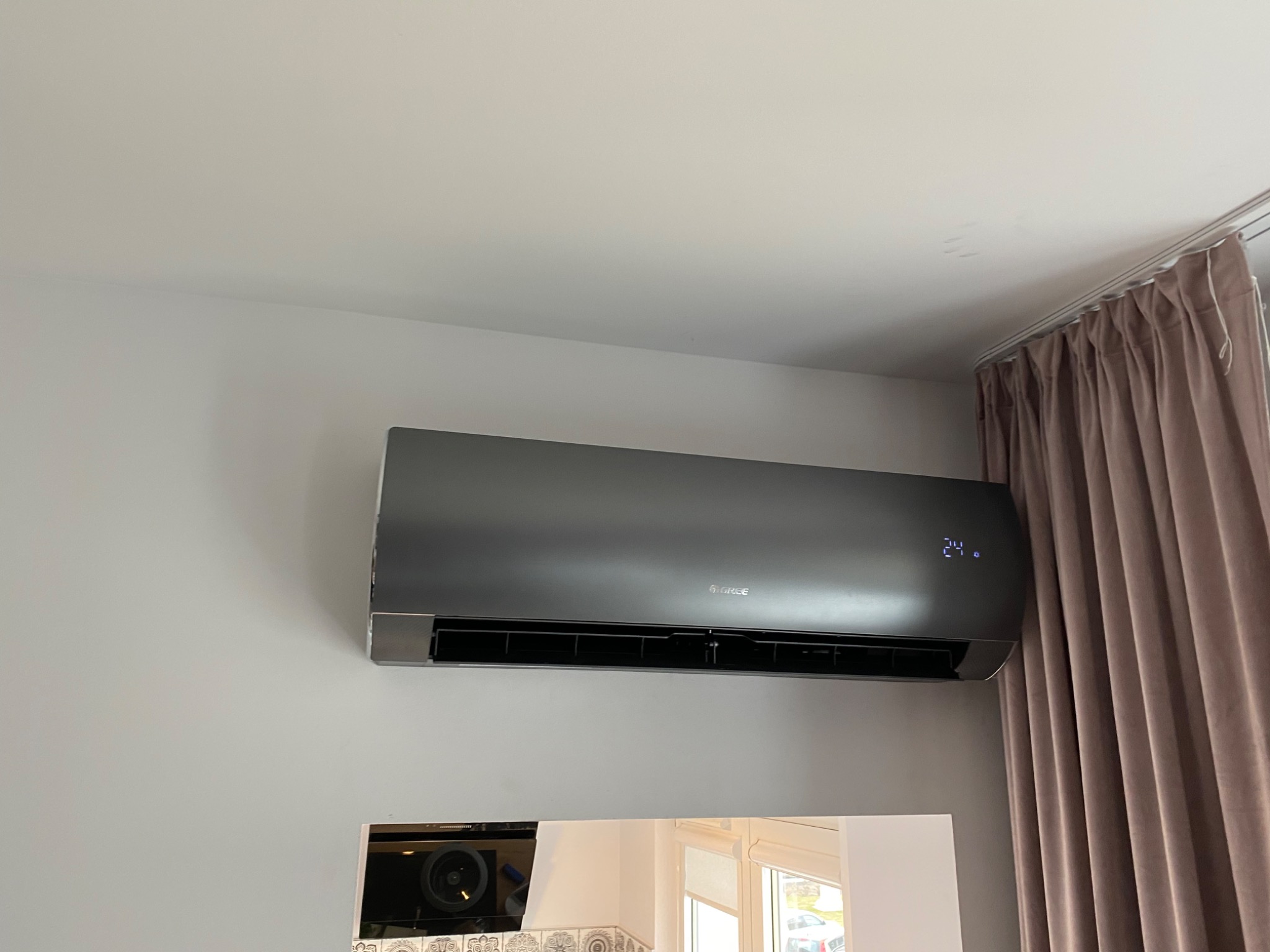 Szary klimatyzator marki Gree zamontowany na jasnej ścianie, z widocznym wyświetlaczem temperatury 24 stopnie, obok zasłony w odcieniach brązu.