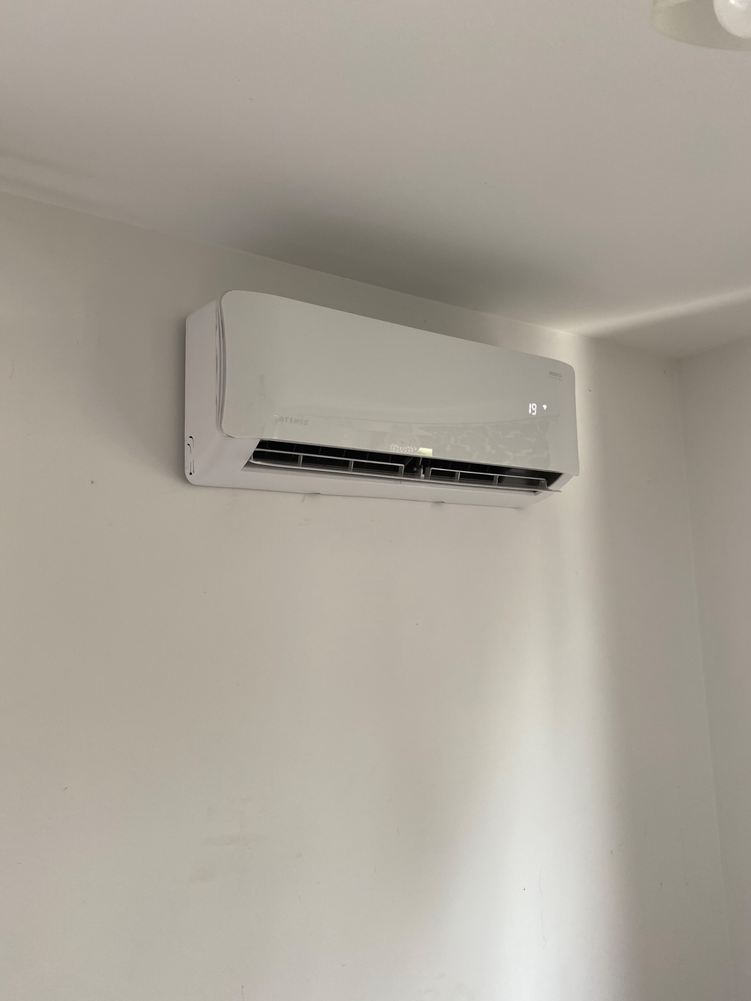 Biały klimatyzator marki Rotenso zamontowany na białej ścianie w rogu pomieszczenia, wyświetlający temperaturę 19 stopni.