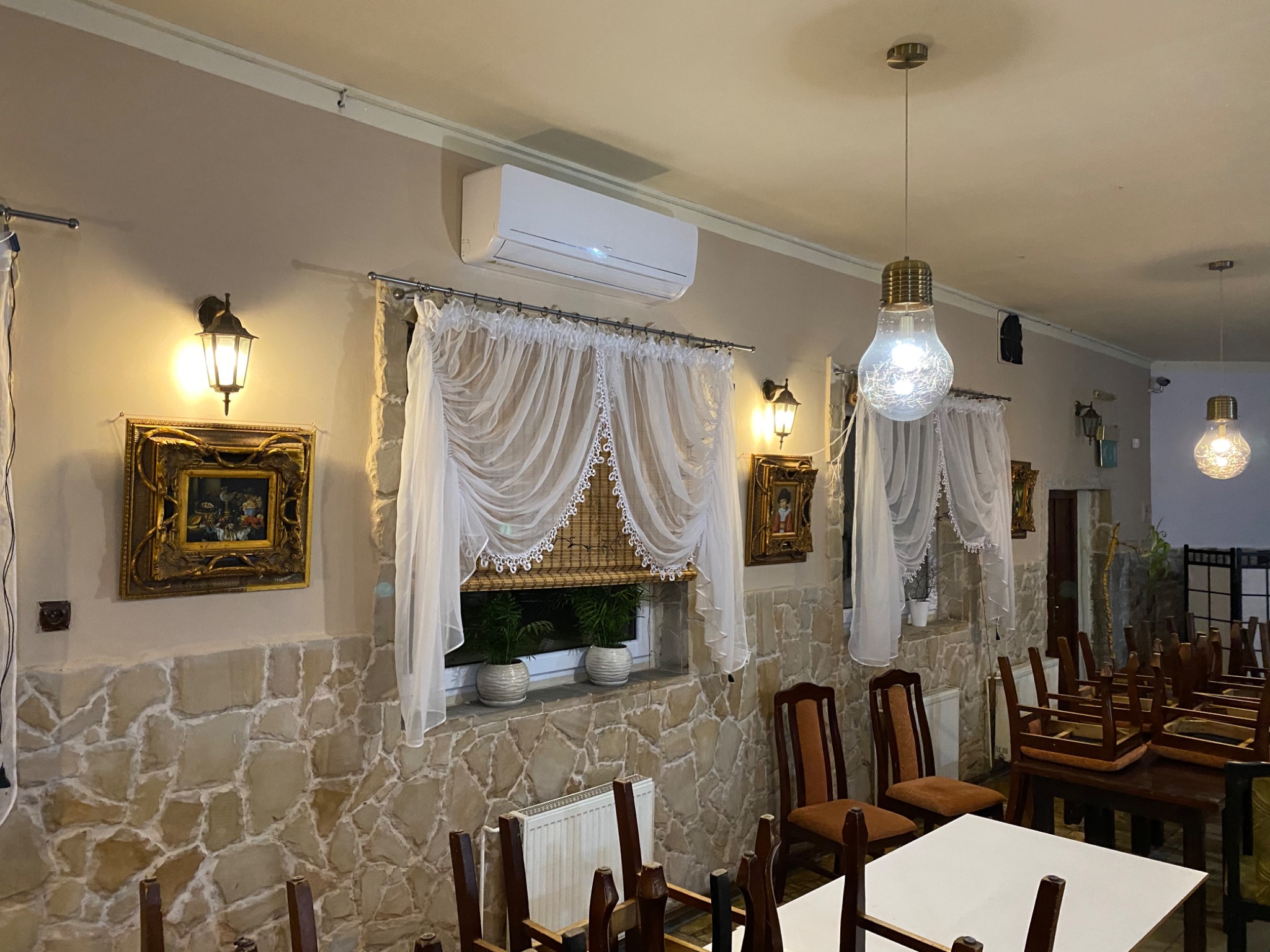 Wnętrze restauracji z klimatyzatorem ściennym, oknami z firanami, kamienną ścianą i dekoracyjnymi lampami w kształcie żarówek.