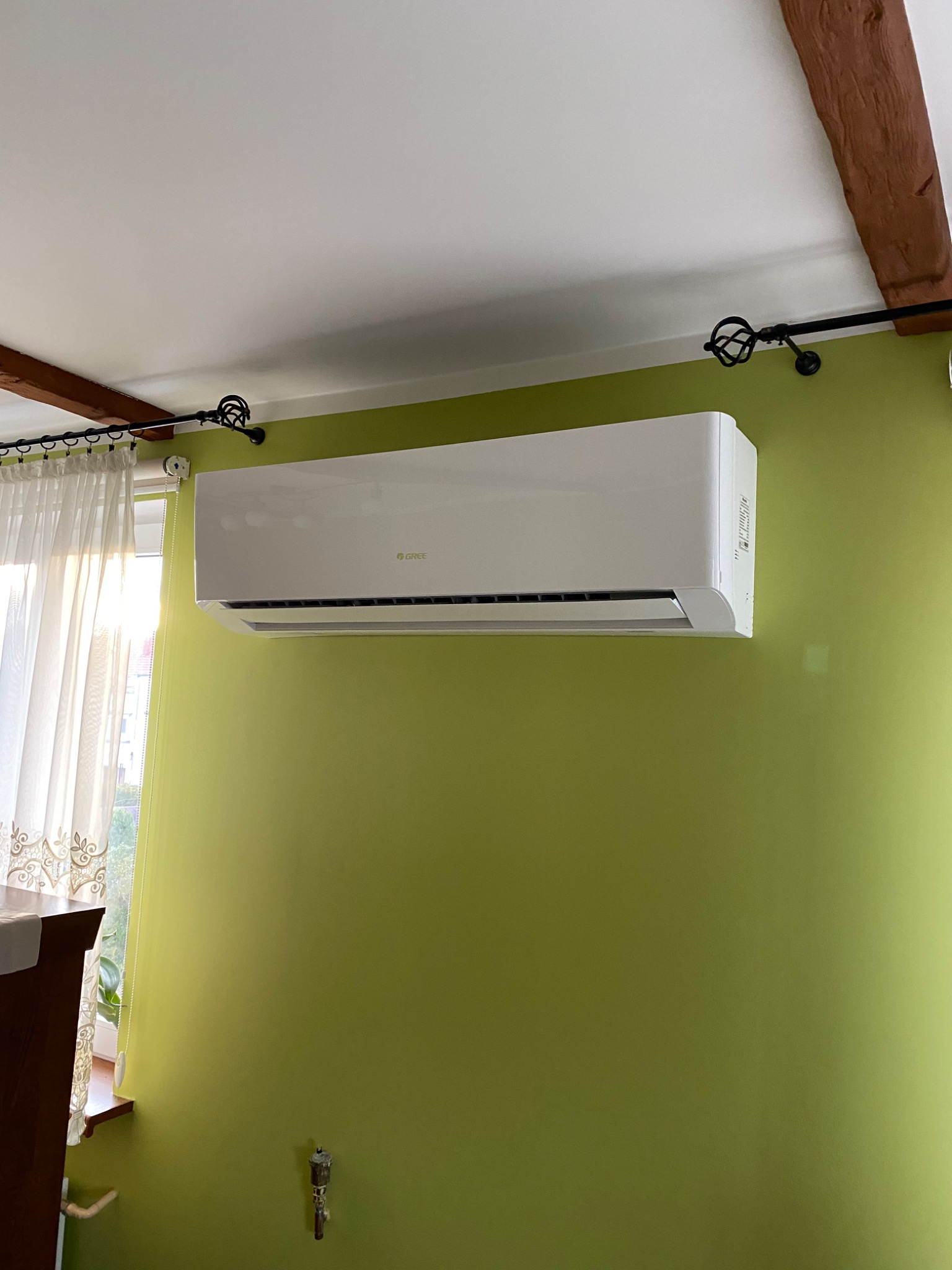 Biały klimatyzator marki Gree zamontowany na zielonej ścianie w pokoju z drewnianą belką stropową i karniszem z ozdobnymi zakończeniami.