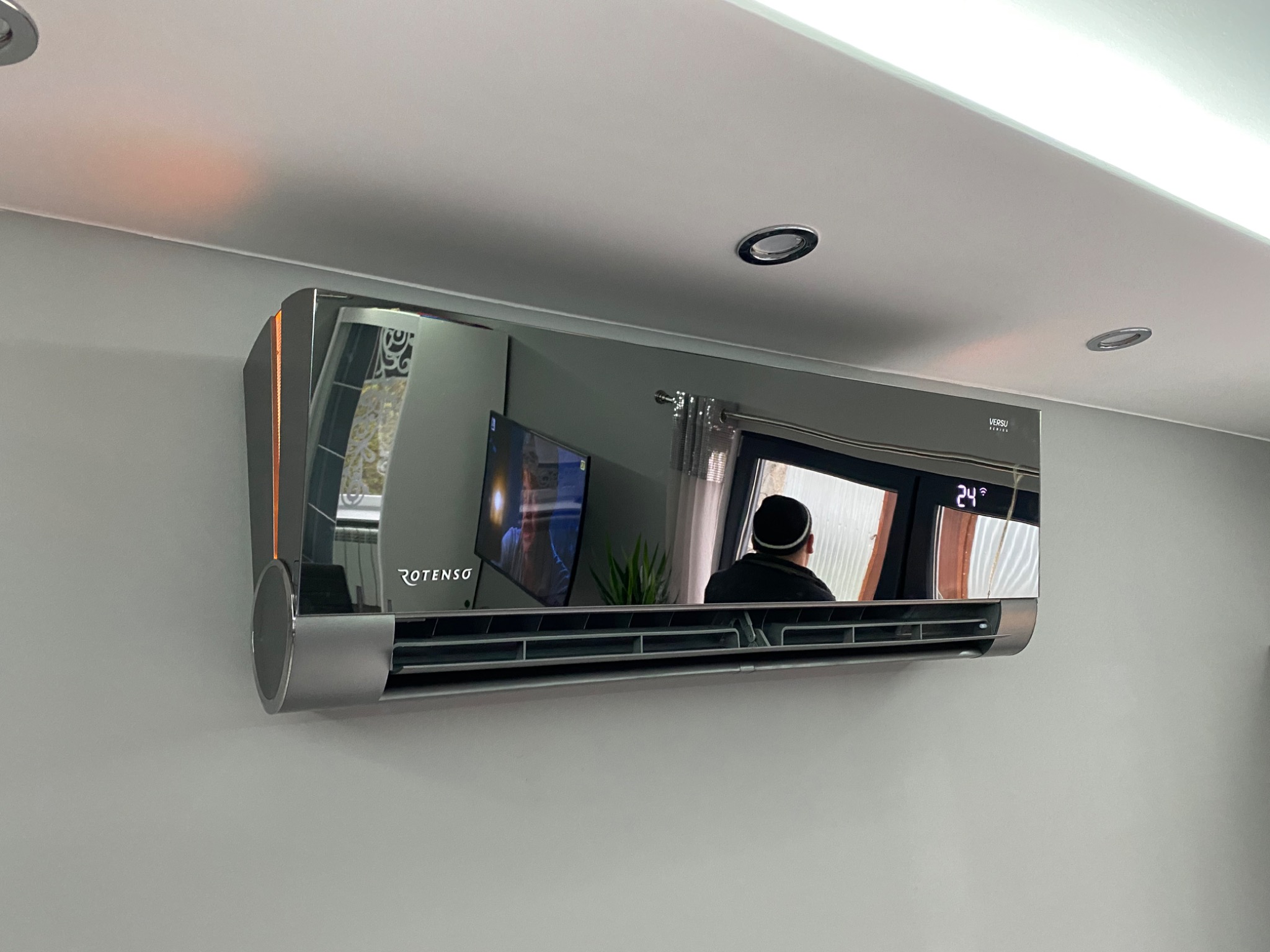 Nowoczesny, srebrny klimatyzator Rotenso zamontowany na ścianie, z widocznym odbiciem wnętrza pokoju, w tym telewizora i osoby w czapce.