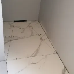 Ułożone białe płytki podłogowe z szarym marmurkowym wzorem w małym pomieszczeniu, widoczna poziomica i krzyżyki dystansowe.