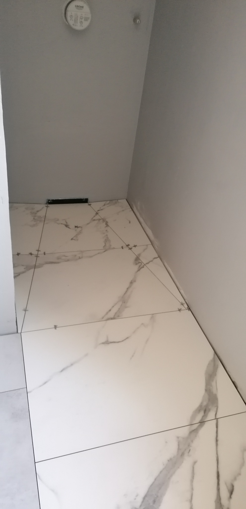 Ułożone białe płytki podłogowe z szarym marmurkowym wzorem w małym pomieszczeniu, widoczna poziomica i krzyżyki dystansowe.