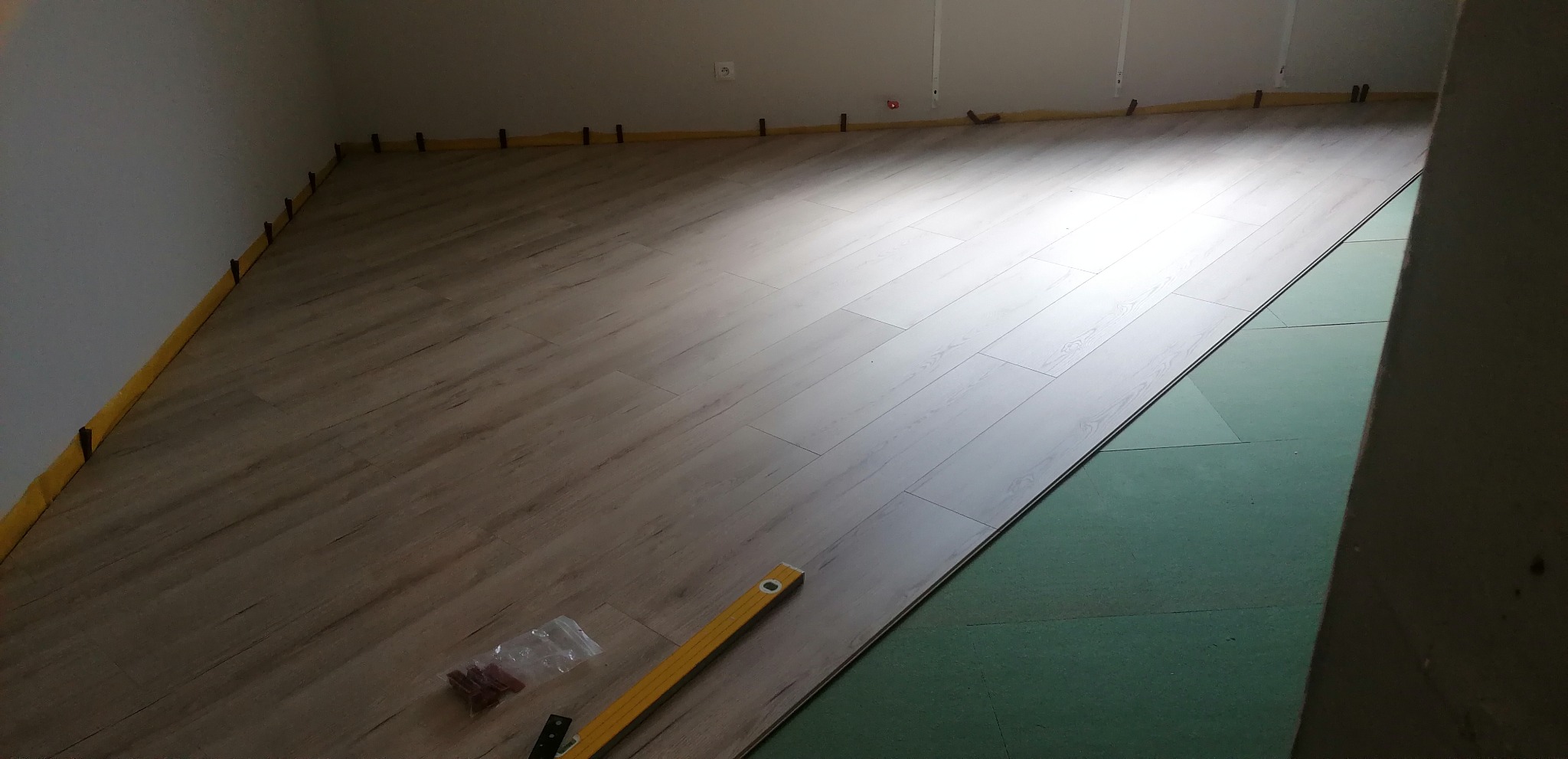 Układanie jasnych paneli podłogowych z widoczną poziomicą i elementami mocującymi przy ścianach.