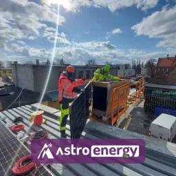 Montaż paneli słonecznych na dachu budynku; jeden z instalatorów w pomarańczowej kurtce pokazuje znak zwycięstwa, drugi w żółtej kurtce rozpakowuje panel z pudełka na podnośniku, na dachu widoczne...