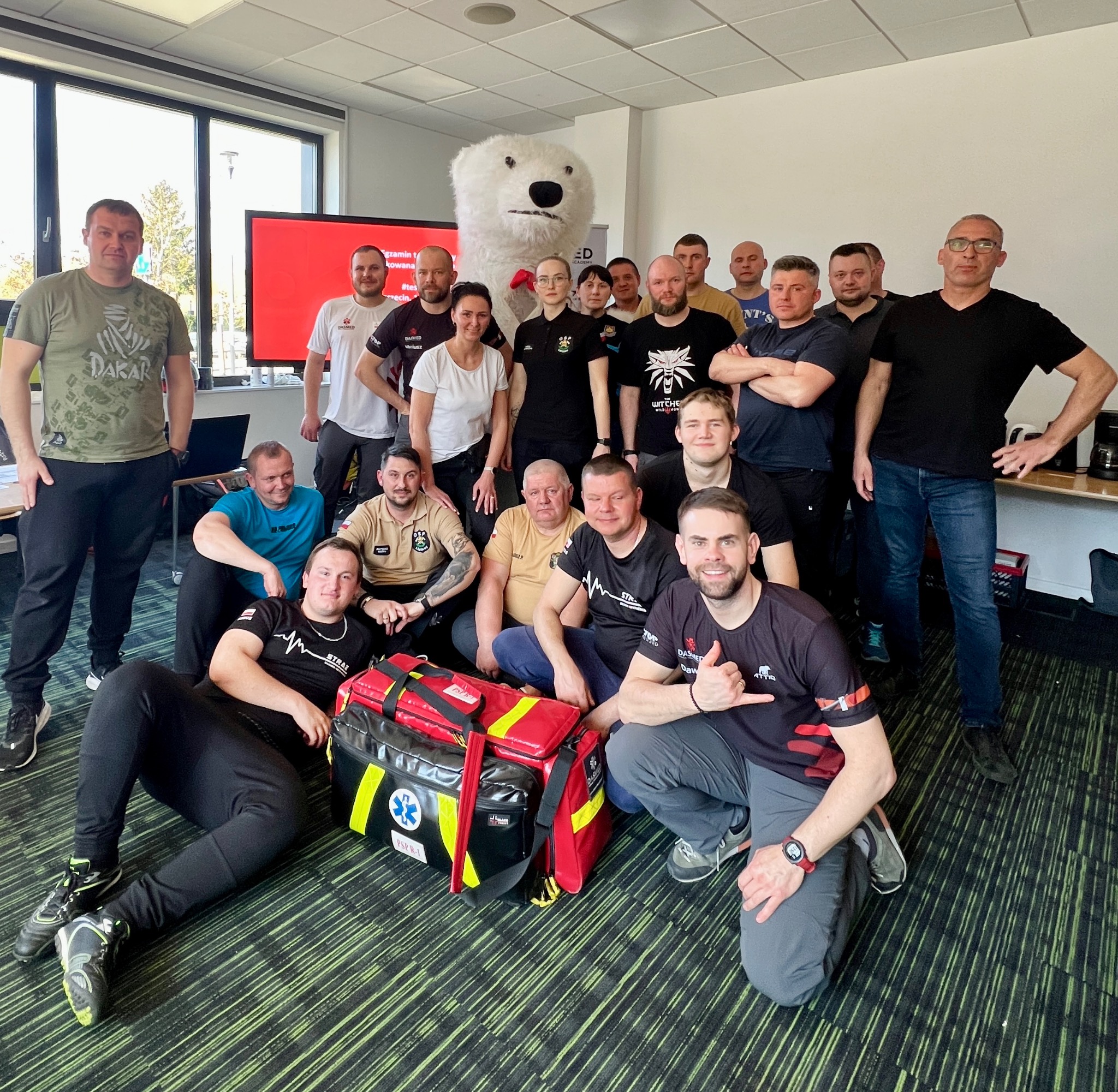 Grupa osób po szkoleniu z pierwszej pomocy, z maskotką misia i torbą medyczną PSPR-1 na podłodze, w jasnym pomieszczeniu z dużym oknem.
