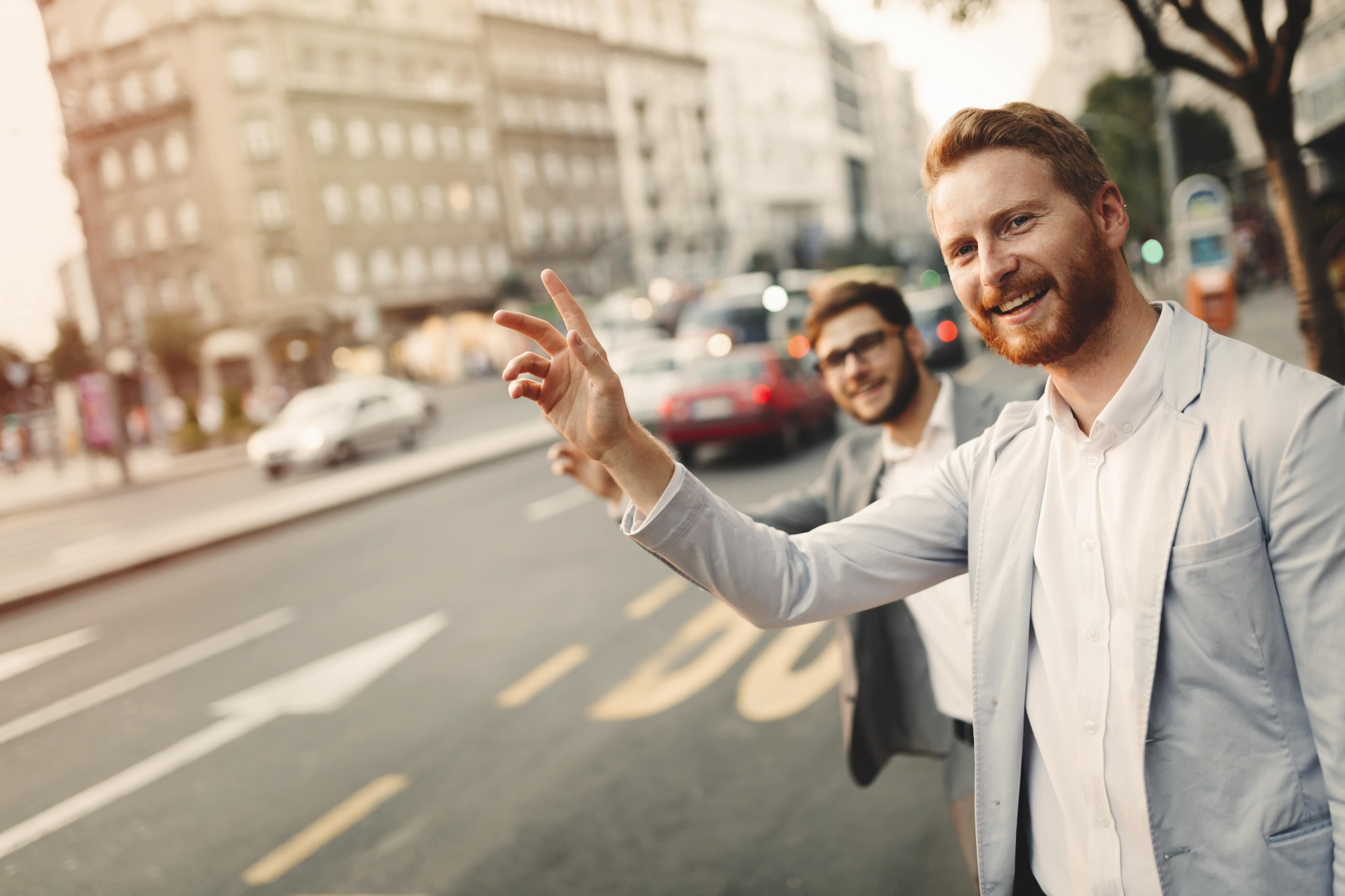 Dwóch eleganckich mężczyzn na ulicy w mieście, jeden z nich macha ręką, próbując zatrzymać taksówkę na tle miejskiego krajobrazu.