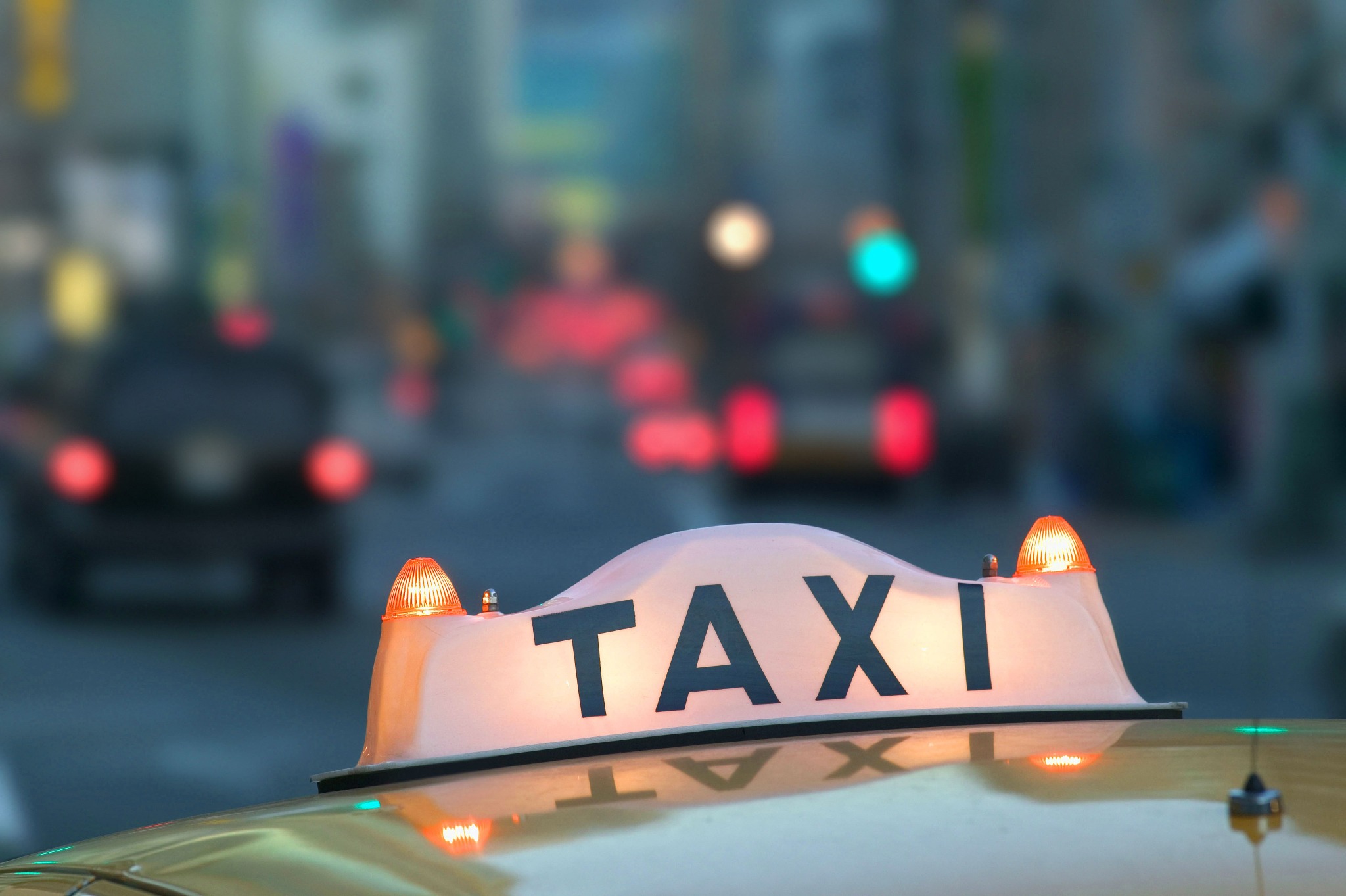 Podświetlony znak TAXI na dachu żółtej taksówki, w tle rozmyte światła miasta i inne pojazdy.