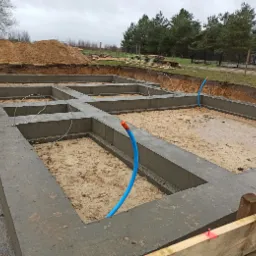 Wylane betonowe fundamenty z wystającymi rurami instalacyjnymi na piaszczystym podłożu, widoczny wykop i pryzma ziemi w tle.