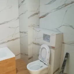 Nowoczesna łazienka z białymi płytkami imitującymi marmur, podwieszana toaleta z otwartą klapą i drewnianą obudową wanny.