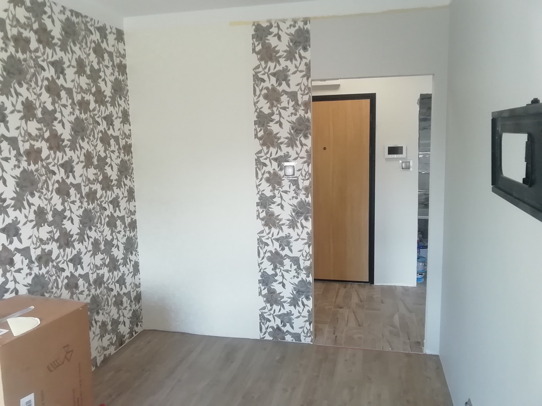 Wnętrze mieszkania w trakcie odświeżania: fragment ściany z tapetą w kwiaty sąsiadujący z gładką, białą ścianą, częściowo widoczna kartonowa skrzynka, podłoga wyłożona panelami w odcieniach szarości.
