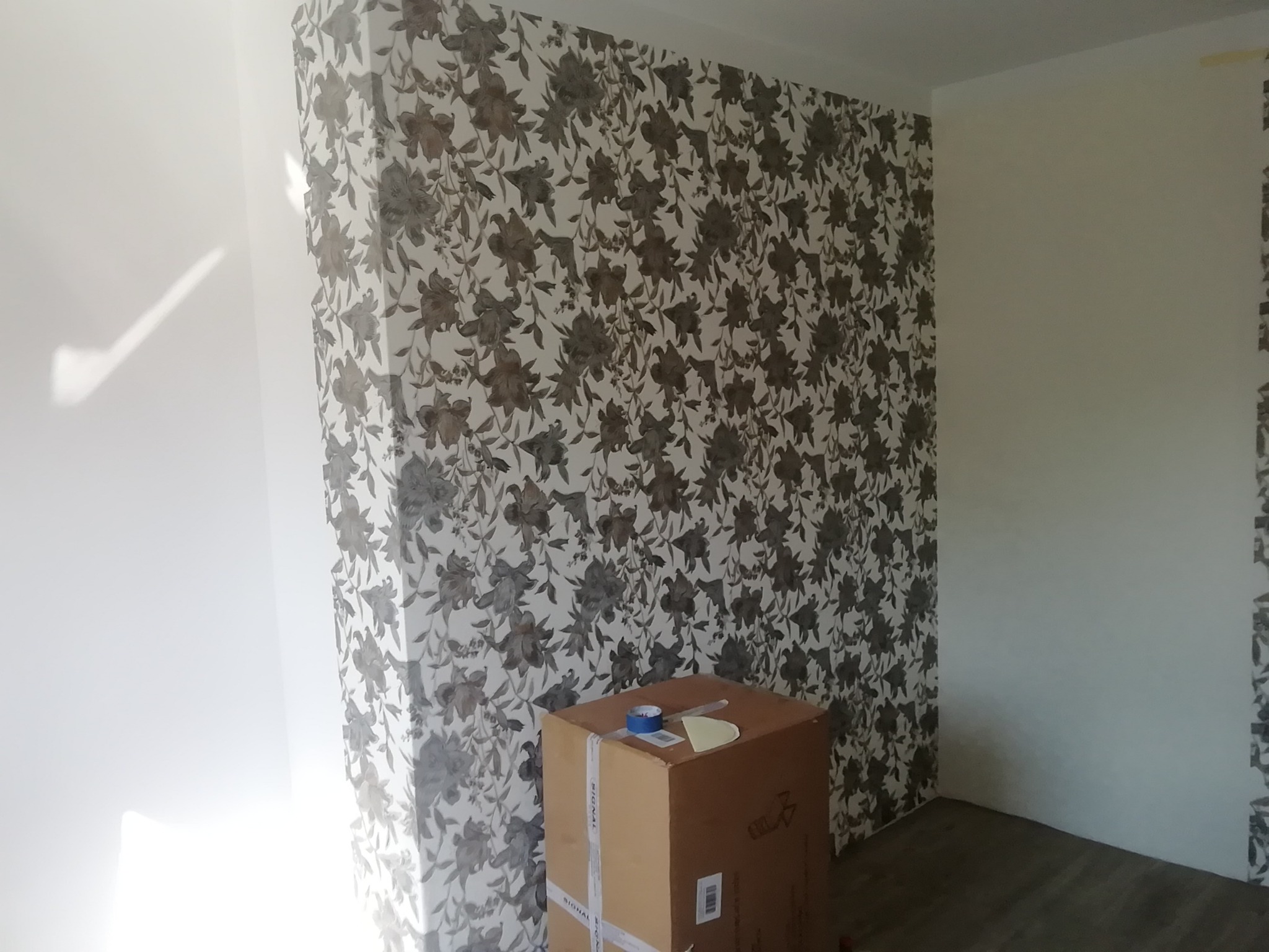 Ściana po położeniu tapety w kwiatowy wzór, częściowo zakryta kartonem z taśmą i nożykiem, widoczny fragment podłogi.