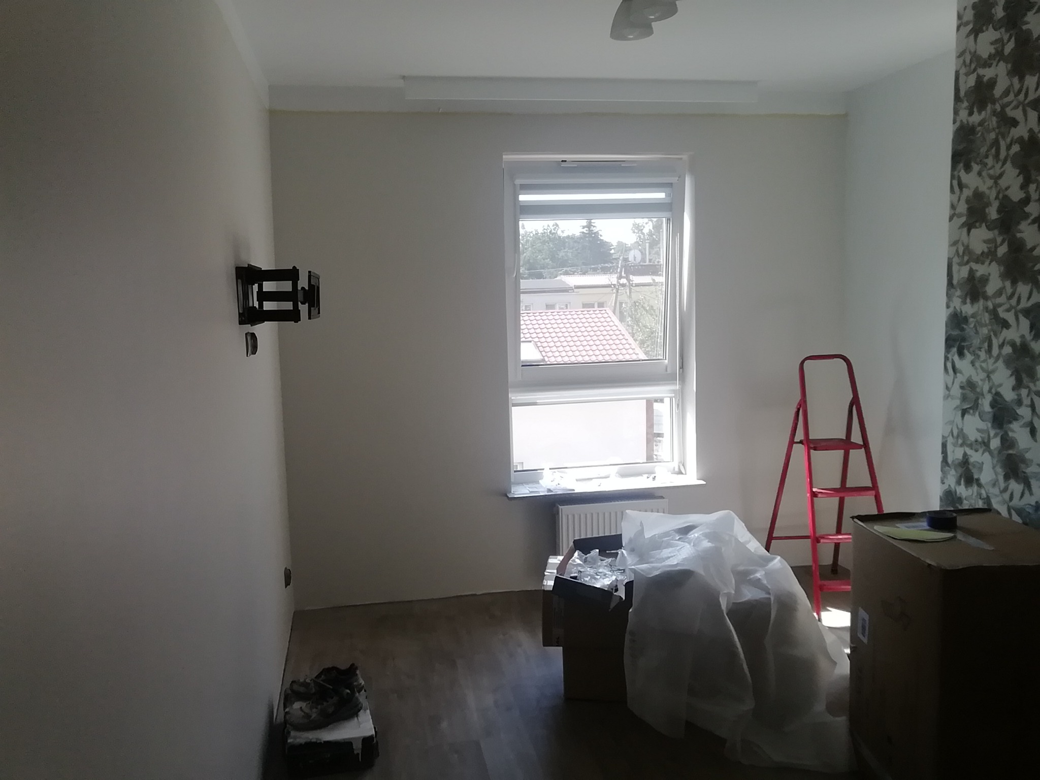 Pokój w trakcie odświeżania: widoczny uchwyt na telewizor na ścianie, okno z roletą, grzejnik, kartony z folią ochronną, czerwona drabina i fragment zasłony w kwiaty.